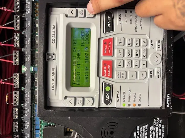 Technician checking a fire alarm control panel showing a heat detector trouble signal